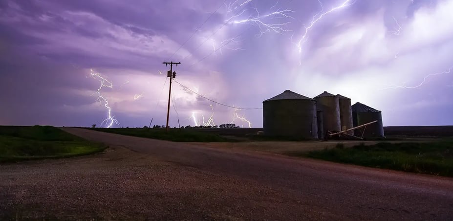 Time lapse photo shows lightning striking in the background, with a utility pole and municipal buildings in the foreground on a dirt road.